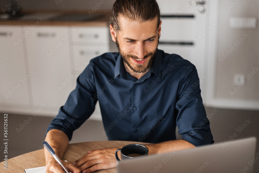 A man smiles, works remotely at a computer. A young man writes information in a notebook and looks into a laptop. A man works at home. View from the top A man smiles, works remotely at a computer. A young man writes information in a notebook and looks into a laptop. A man works at home. View from the top