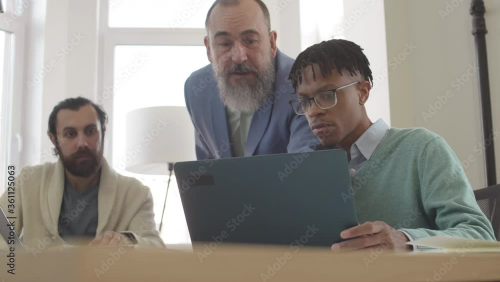 Medium shot of old Caucasian businessman is standing behind two diverse men sitting at desk, working on computers and looking at laptop screen while talking to African man Medium shot of old Caucasian businessman is standing behind two diverse men sitting at desk, working on computers and looking at laptop screen while talking to African man