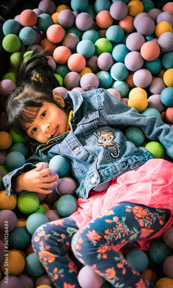 Little asian girl having fun in ball pit with colorful balls. Child playing on indoor playground. Kid jumping in ball pool.
