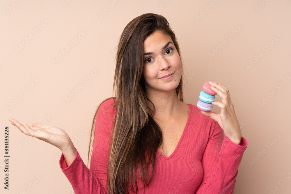 Teenager girl over isolated background holding colorful French macarons and making doubts gesture Teenager girl over isolated background holding colorful French macarons and making doubts gesture