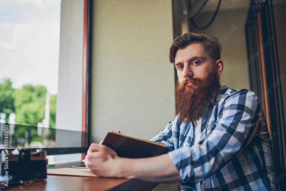 Half length portrait of skilled bearded student with textbook in hand looking at camera while sitting in coffee shop and preparing for upcoming exams.Publicity area for your advertising text message Half length portrait of skilled bearded student with textbook in hand looking at camera while sitting in coffee shop and preparing for upcoming exams.Publicity area for your advertising text message