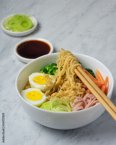 Ramen noodles soup in white bowl with eggs, scallion, chives, broccoli, carrot, ham, noodles wrapped in chopsticks, copy space