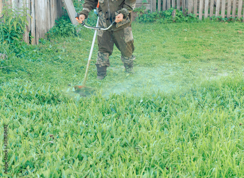 Asian man in a mosquito suit with a professional brush cutter trimmer mows the grass in the yard. green lawn, old fence of the village house. Sunny summer weather. cutting  lawn mower.