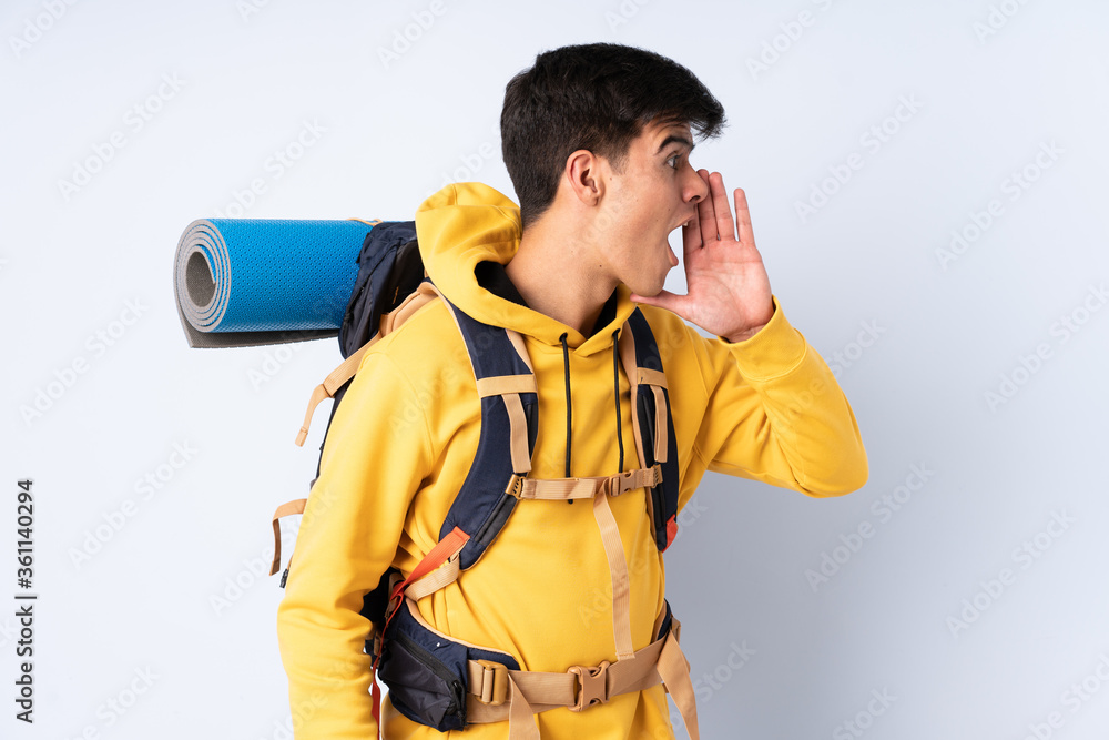 Young mountaineer man with a big backpack over isolated blue background shouting with mouth wide open Young mountaineer man with a big backpack over isolated blue background shouting with mouth wide open