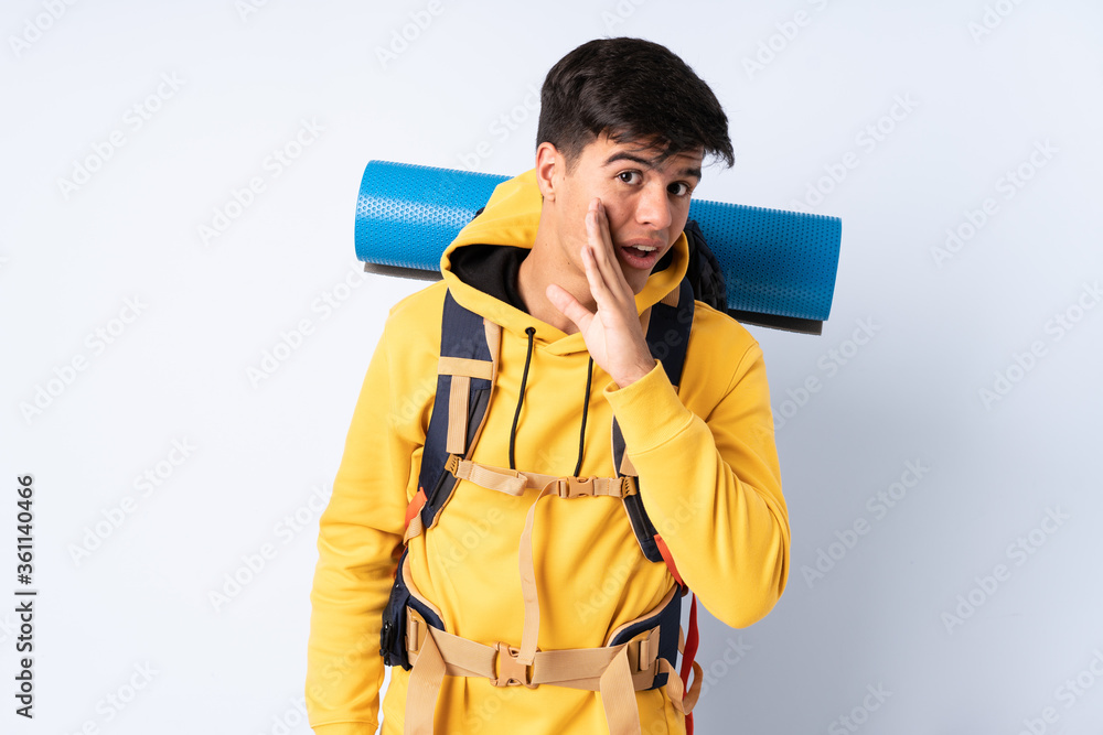 Young mountaineer man with a big backpack over isolated blue background whispering something Young mountaineer man with a big backpack over isolated blue background whispering something