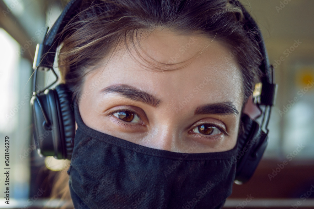 A young girl in a protective mask and headphones looks at the camera. A brunette in a protective mask listens to music and rides the train A young girl in a protective mask and headphones looks at the camera. A brunette in a protective mask listens to music and rides the train