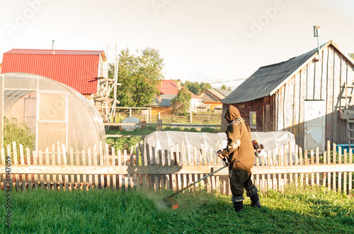 Asian man in a mosquito suit with a professional brush cutter trimmer mows the grass in the yard. green lawn, old fence of the village house. Sunny summer weather. cutting lawn mower.