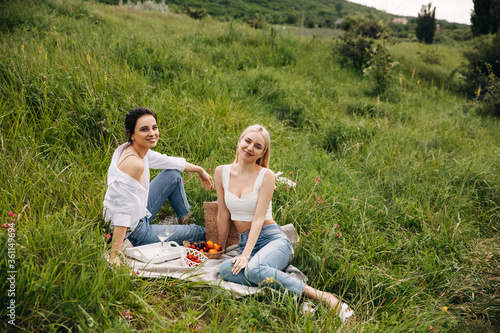 Two young women smiling, sitting on a blanket on fresh green grass at a picnic, outdoors, in mountains.