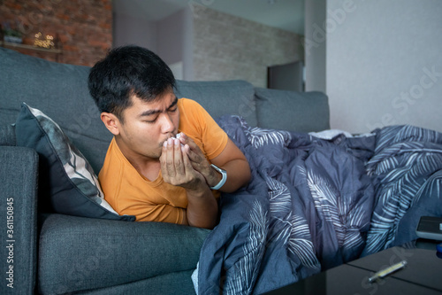 Asian man is ill and coughs on the sofa at home and he uses a thermometer to check the temperature during the coronavirus epidemic.