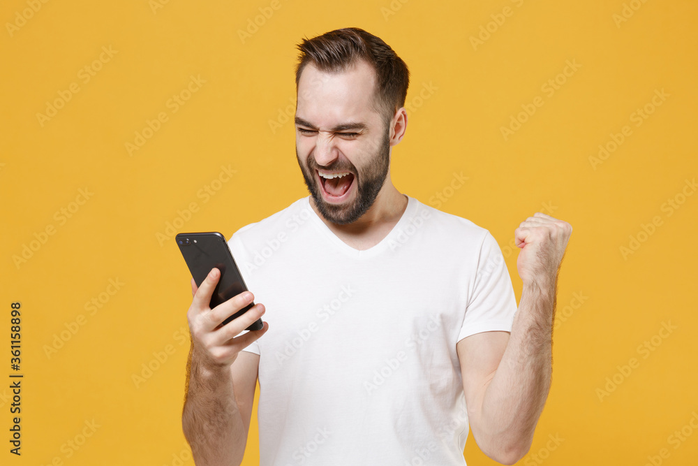 Happy young bearded man guy in white casual t-shirt posing isolated on yellow wall background studio portrait. People lifestyle concept. Mock up copy space. Using mobile phone, doing winner gesture. Happy young bearded man guy in white casual t-shirt posing isolated on yellow wall background studio portrait. People lifestyle concept. Mock up copy space. Using mobile phone, doing winner gesture.