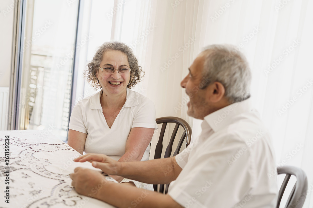 Old aged, Turkish, happy senior couple enjoying time together, looking to each other, laughing, sitting in living room on chair.	