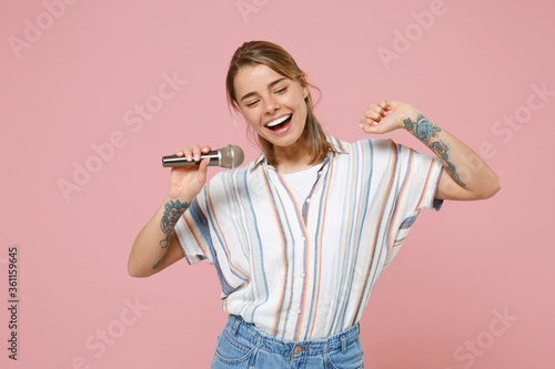 Cheerful young blonde woman girl in casual striped shirt posing isolated on pink background studio portrait. People sincere emotions lifestyle concept. Mock up copy space. Sing song in microphone.