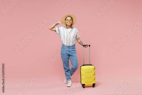 Cheerful traveler girl in casual striped shirt eyeglasses hat isolated on pink background. Passenger traveling abroad to travel on weekends getaway. Air flight journey concept. Hold yellow suitcase.