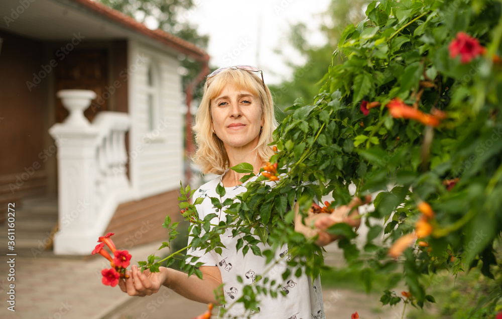 Beautiful woman standing in the yard near the bush with flowers and posing for the camera. Adult blonde woman resting in a country house, standing near a beautiful bush and looking at the camera