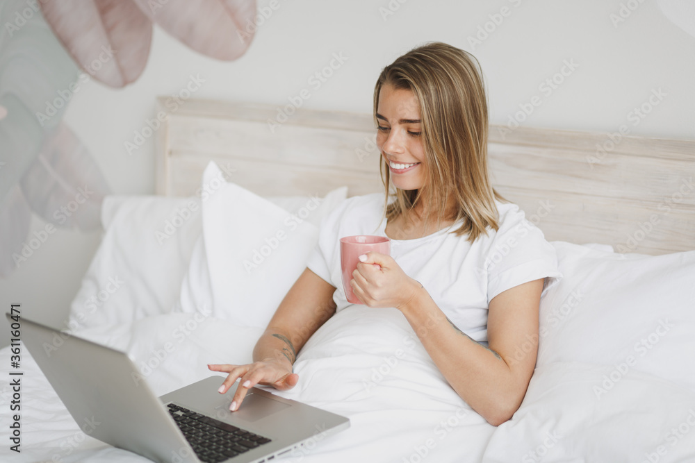 Smiling young woman girl lying in bed with white sheet pillow blanket spending time in bedroom at home. Rest relax good mood lifestyle concept. Working on laptop pc computer hold cup of coffee or tea. Smiling young woman girl lying in bed with white sheet pillow blanket spending time in bedroom at home. Rest relax good mood lifestyle concept. Working on laptop pc computer hold cup of coffee or tea.