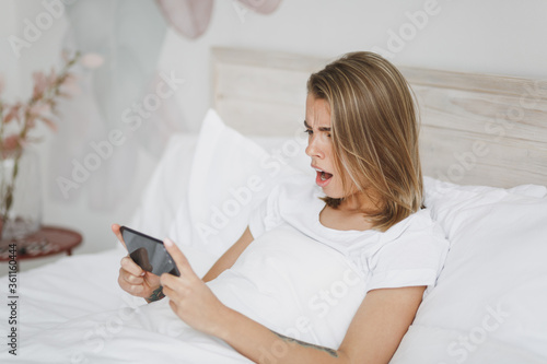 Shocked girl lying in bed with white sheet pillow blanket spending time in bedroom at home. Rest relax good mood lifestyle concept. Mock up copy space. Using mobile phone, play game, watching video.