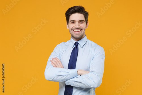 Smiling young business man in classic blue shirt tie posing isolated on yellow wall background studio portrait. Achievement career wealth business concept. Mock up copy space. Holding hands crossed.