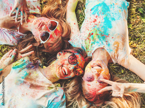 Three happy beautiful girls making party at Holi colors festival in summer time.Young smiling women friends having fun after music event at sunset.Models lying on the grass in sunglasses.