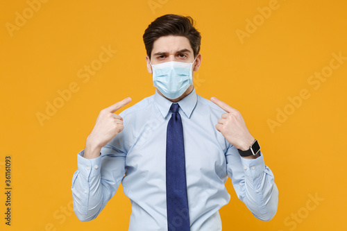 Young business man in classic blue shirt tie isolated on yellow background studio. Epidemic pandemic coronavirus 2019-ncov sars covid-19 flu virus concept. Pointing index fingers on sterile face mask.