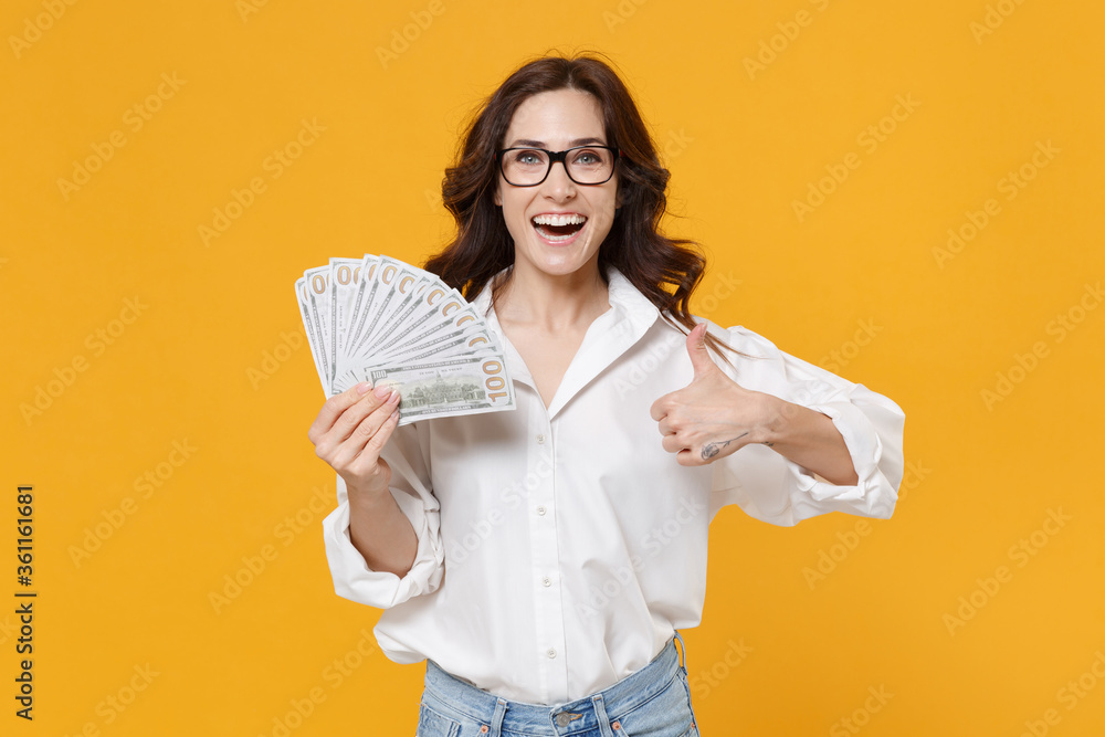Funny young brunette business woman in white shirt glasses isolated on yellow wall background. Achievement career wealth business concept. Hold fan of cash money in dollar banknotes, showing thumb up. Funny young brunette business woman in white shirt glasses isolated on yellow wall background. Achievement career wealth business concept. Hold fan of cash money in dollar banknotes, showing thumb up.