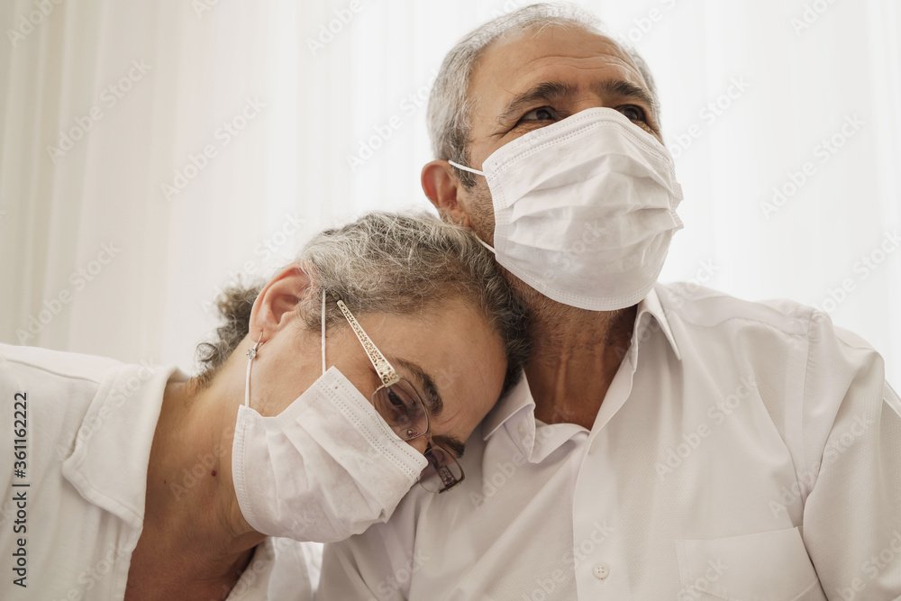 Sad looking old couple wearing masks. Wife puts her head on her husband's shoulder