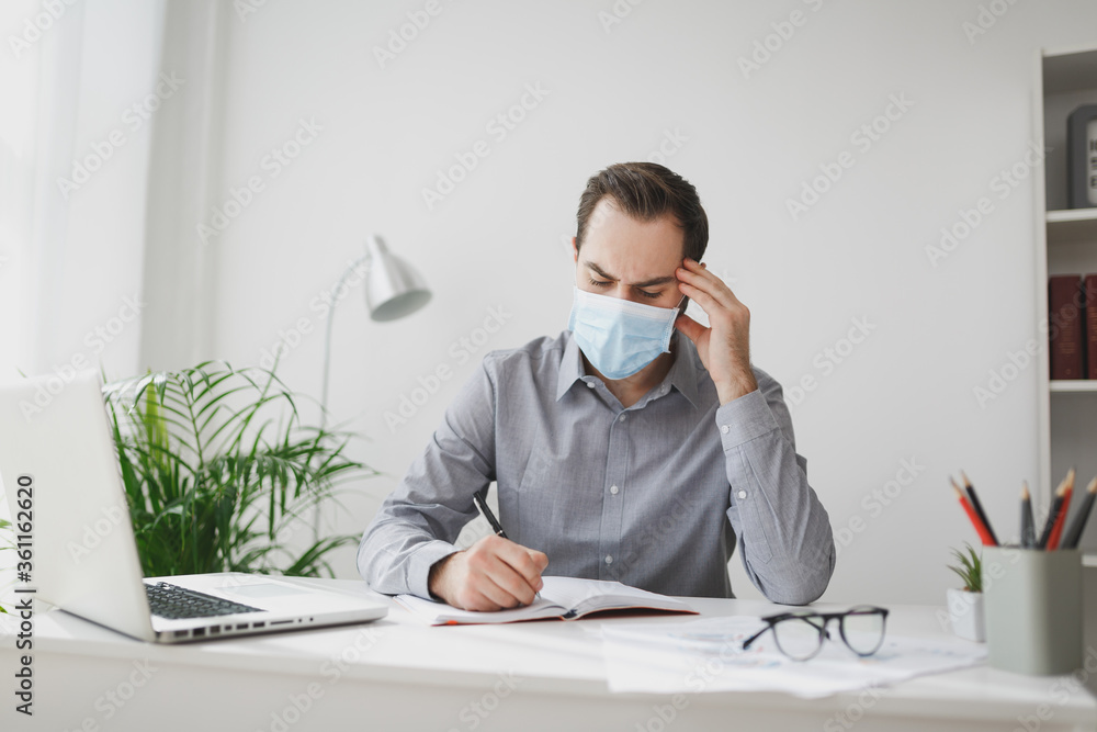 Young business man in gray shirt sterile face mask sit at desk work on laptop in light office on white wall background. Achievement business career concept. Writing notes in notebook put hand on head. Young business man in gray shirt sterile face mask sit at desk work on laptop in light office on white wall background. Achievement business career concept. Writing notes in notebook put hand on head.
