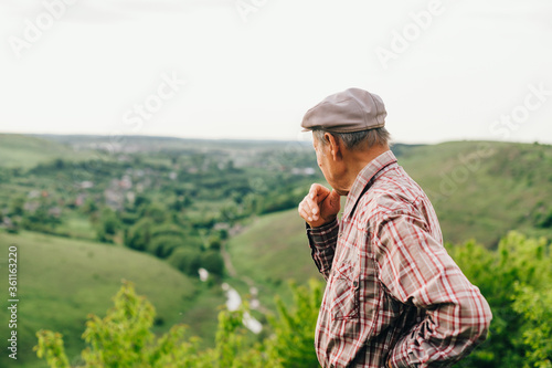 A pensive old retired man stands on top of a mountain and looks at a beautiful landscape. Retired man traveling. Old grandfather looks at the beautiful landscape from the mountain and thinks