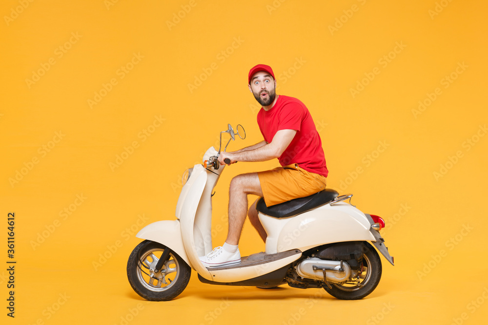 Fototapeta premium Delivery man in red cap t-shirt uniform driving moped motorbike scooter isolated on yellow background studio Guy employee working courier Service quarantine pandemic coronavirus virus covid-19 concept