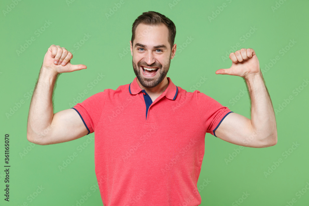 Fototapeta premium Funny young bearded man guy in casual red pink t-shirt posing isolated on green background studio portrait. People sincere emotions lifestyle concept. Mock up copy space. Pointing thumbs on himself.