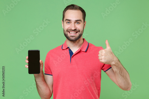 Smiling young bearded man guy in casual red pink t-shirt posing isolated on green background. People lifestyle concept. Mock up copy space. Hold mobile phone with blank empty screen showing thumb up.