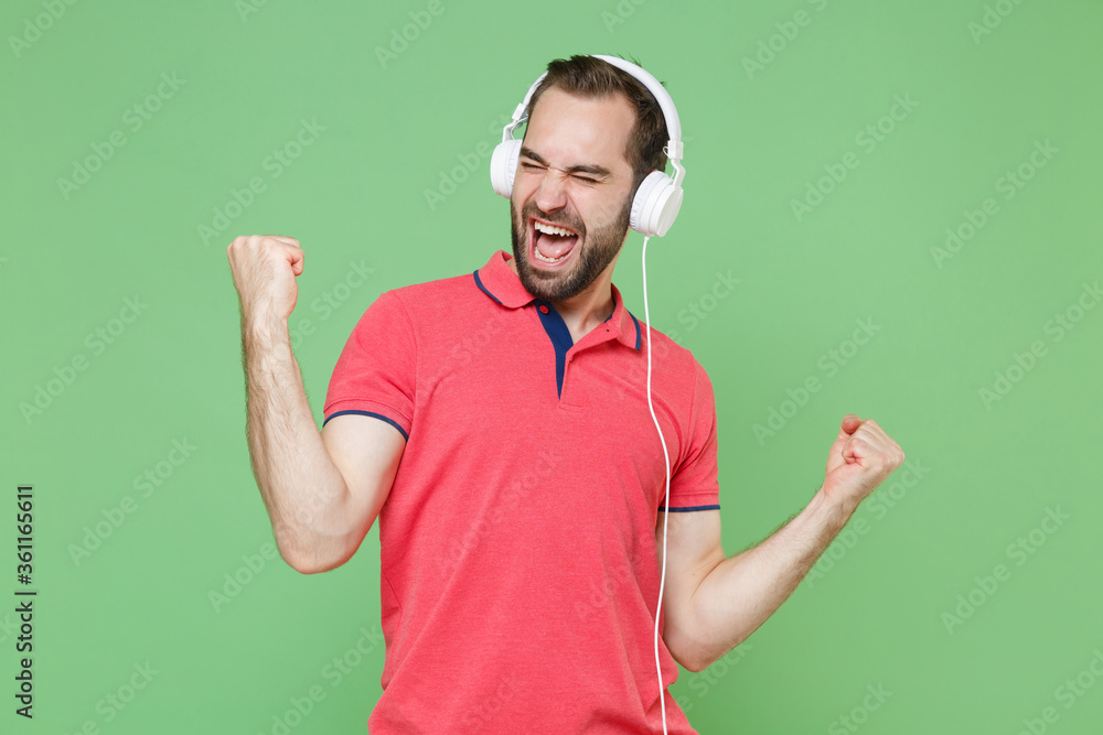 Screaming young bearded man guy in casual red pink t-shirt isolated on green wall background. People lifestyle concept. Listen music with headphones, keeping eyes closed, clenching fists like winner. Screaming young bearded man guy in casual red pink t-shirt isolated on green wall background. People lifestyle concept. Listen music with headphones, keeping eyes closed, clenching fists like winner.