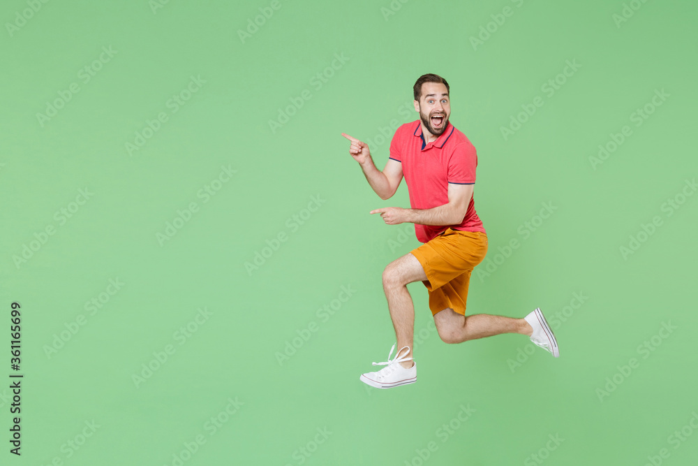 Excited young bearded man guy in casual red pink t-shirt posing isolated on green wall background studio portrait. People lifestyle concept. Mock up copy space. Jumping pointing index fingers aside. Excited young bearded man guy in casual red pink t-shirt posing isolated on green wall background studio portrait. People lifestyle concept. Mock up copy space. Jumping pointing index fingers aside.