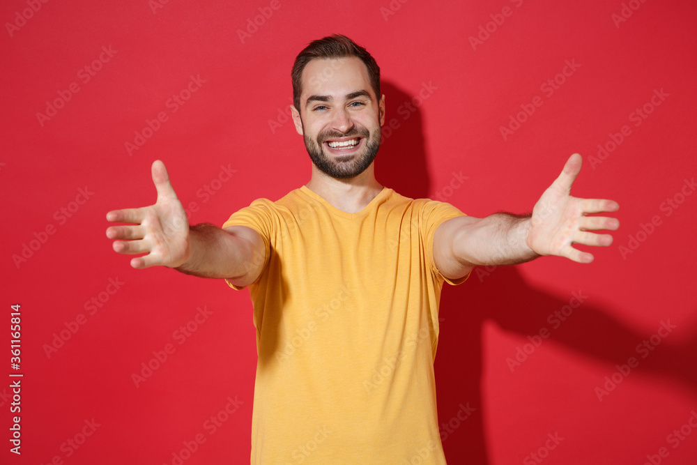 Smiling young bearded man guy in casual yellow t-shirt posing isolated on red wall background studio portrait. People lifestyle concept. Mock up copy space. Standing with outstretched hands for hugs. Smiling young bearded man guy in casual yellow t-shirt posing isolated on red wall background studio portrait. People lifestyle concept. Mock up copy space. Standing with outstretched hands for hugs.