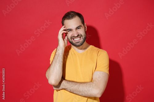 Displeased puzzled young bearded man guy in casual yellow t-shirt posing isolated on red background studio portrait. People sincere emotions lifestyle concept. Mock up copy space. Put hand on head.