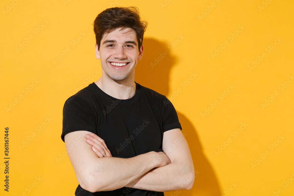 Smiling young man guy 20s wearing casual black t-shirt posing isolated on yellow wall background studio portrait. People sincere emotions lifestyle concept. Mock up copy space. Holding hands crossed. Smiling young man guy 20s wearing casual black t-shirt posing isolated on yellow wall background studio portrait. People sincere emotions lifestyle concept. Mock up copy space. Holding hands crossed.