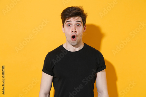 Shocked young man guy 20s wearing casual black t-shirt posing isolated on yellow wall background studio portrait. People sincere emotions lifestyle concept. Mock up copy space. Keeping mouth open. © ViDi Studio