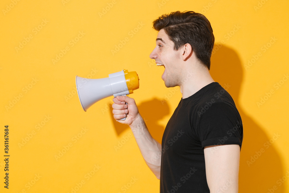 Side veiw of shocked young man guy in casual black t-shirt posing isolated on yellow background studio portrait. People sincere emotions lifestyle concept. Mock up copy space. Screaming in megaphone. Side veiw of shocked young man guy in casual black t-shirt posing isolated on yellow background studio portrait. People sincere emotions lifestyle concept. Mock up copy space. Screaming in megaphone.
