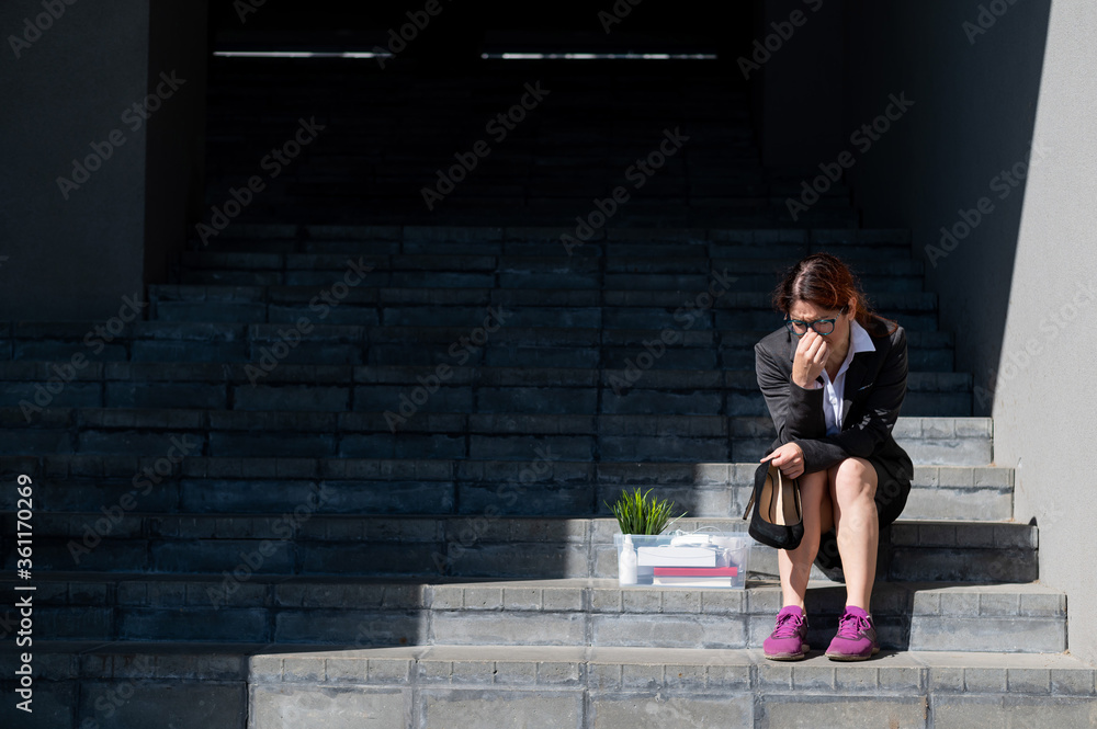 Depressed woman dressed in medical mask is fired and is sitting on the stairs with a box of personal belongings. Female office worker in suit and sneakers outdoors. Unemployment in the economic crisis