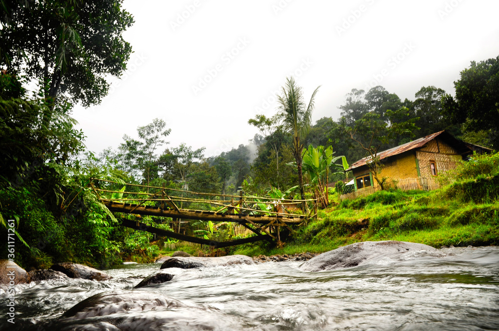 Tropical landscape with rocky river under the wooden bridge and typical rural Indonesian house in the Jungle of west Java Tropical landscape with rocky river under the wooden bridge and typical rural Indonesian house in the Jungle of west Java