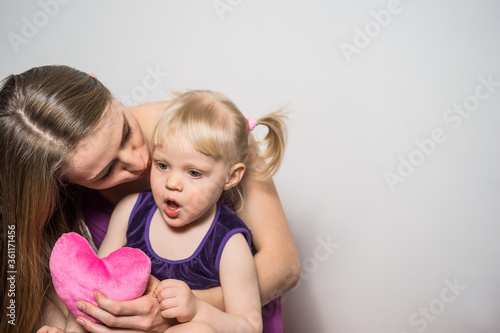 Mother kisses her daughter and gives her love and pink plush heart. The daughter opened her mouth in surprise on light gray background