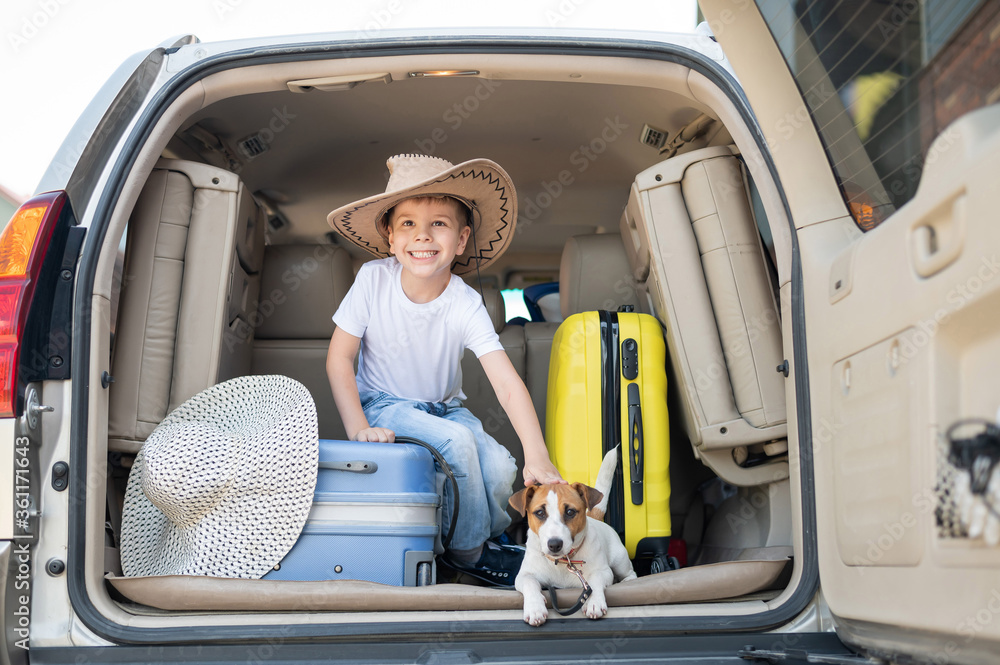 Happy boy in a cowboy hat and puppy jack russell terrier travel by car. A child and a funny little dog are sitting in the trunk and are ready for summer vacation. Independent travel. Best friends.