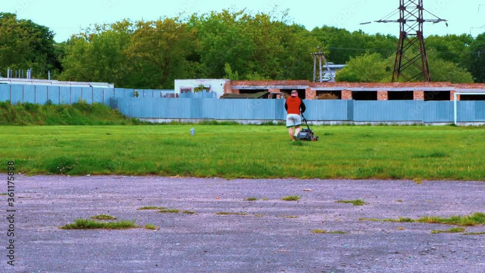 A male handyman mows a grass with a lawn mower in an old abandoned stadium. A male handyman mows a grass with a lawn mower in an old abandoned stadium.