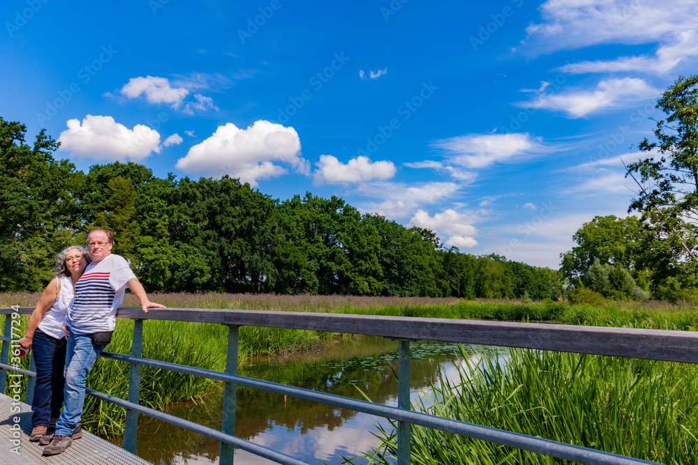 Mature couple, a mexican woman and a dutch man standing on a metal bridge over a stream with trees and green vegetation in the background, sunny spring day in South Limburg, the Netherlands