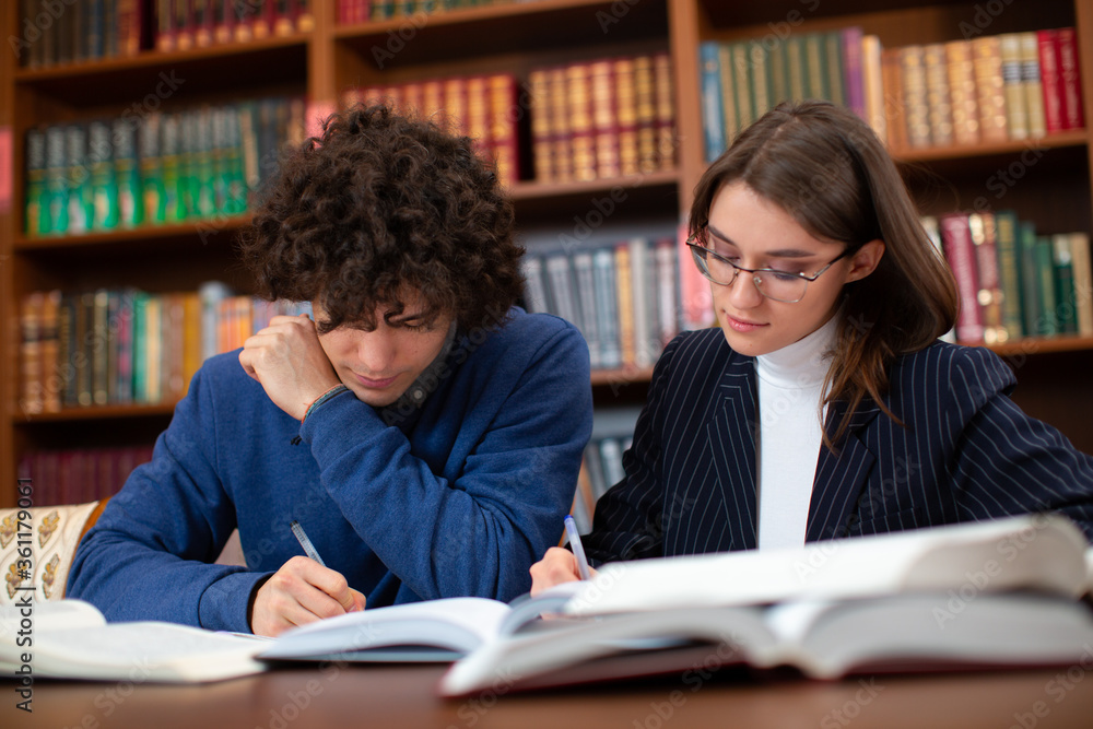 University students, a curly guy in a blue sweater and a brunette girl in glasses in a black jacket doing their homework in the library. Students learn hard.