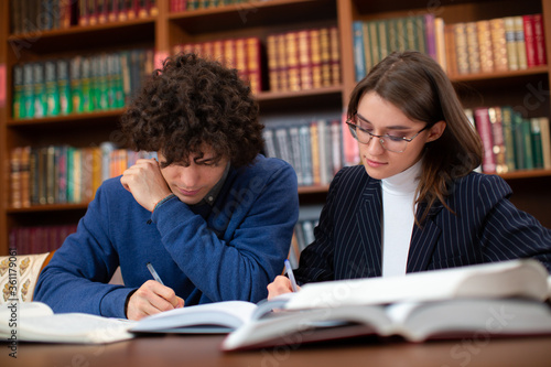 University students, a curly guy in a blue sweater and a brunette girl in glasses in a black jacket doing their homework in the library. Students learn hard.