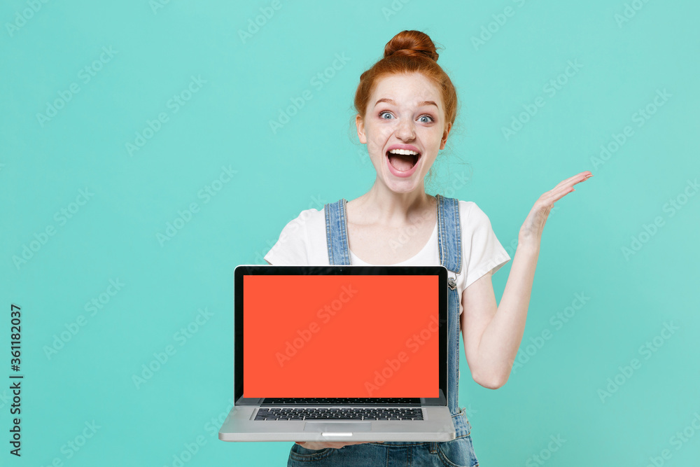 Naklejka premium Excited young readhead girl in casual denim clothes isolated on blue turquoise background. People lifestyle concept. Mock up copy space Hold laptop pc computer with blank empty screen spreading hands.