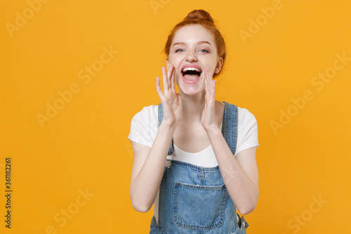 Cheerful young readhead girl in casual denim clothes white t-shirt isolated on yellow background studio portrait. People lifestyle concept. Mock up copy space. Screaming with hands gesture near mouth.