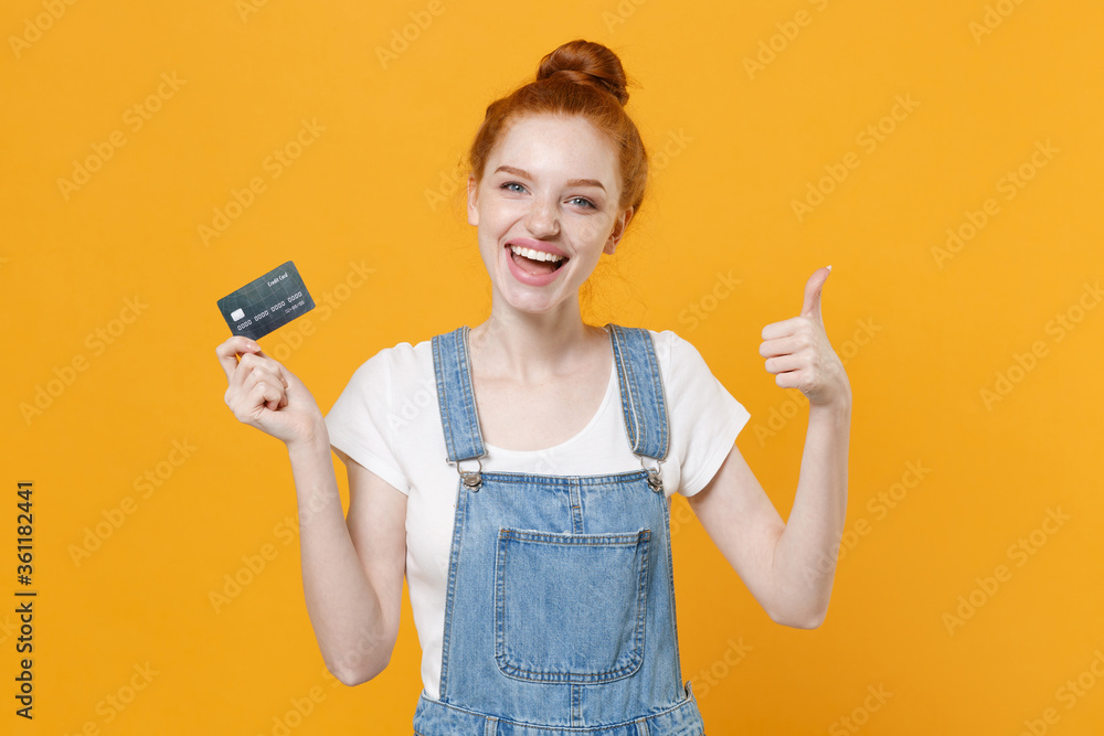 Cheerful young readhead girl in casual denim clothes white t-shirt isolated on yellow background studio portrait. People lifestyle concept. Mock up copy space. Hold credit bank card, showing thumb up. Cheerful young readhead girl in casual denim clothes white t-shirt isolated on yellow background studio portrait. People lifestyle concept. Mock up copy space. Hold credit bank card, showing thumb up.