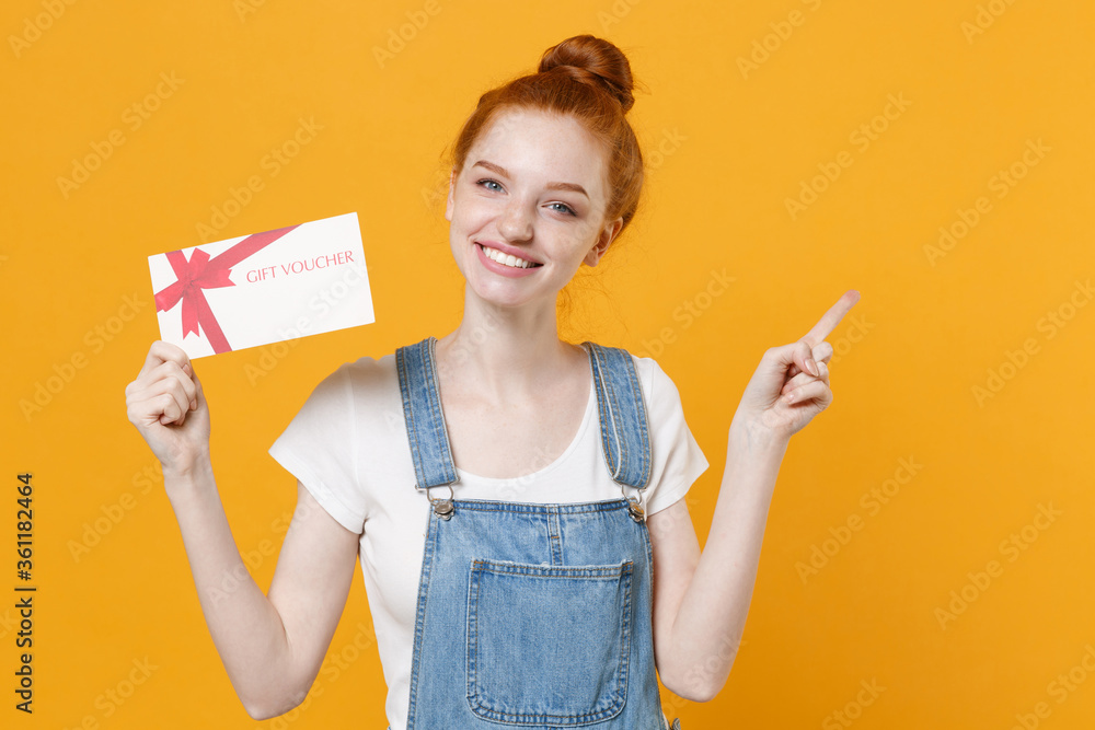 Smiling young readhead girl in casual denim clothes white t-shirt isolated on yellow background. People lifestyle concept. Mock up copy space. Hold gift certificate, pointing index finger aside up. Smiling young readhead girl in casual denim clothes white t-shirt isolated on yellow background. People lifestyle concept. Mock up copy space. Hold gift certificate, pointing index finger aside up.