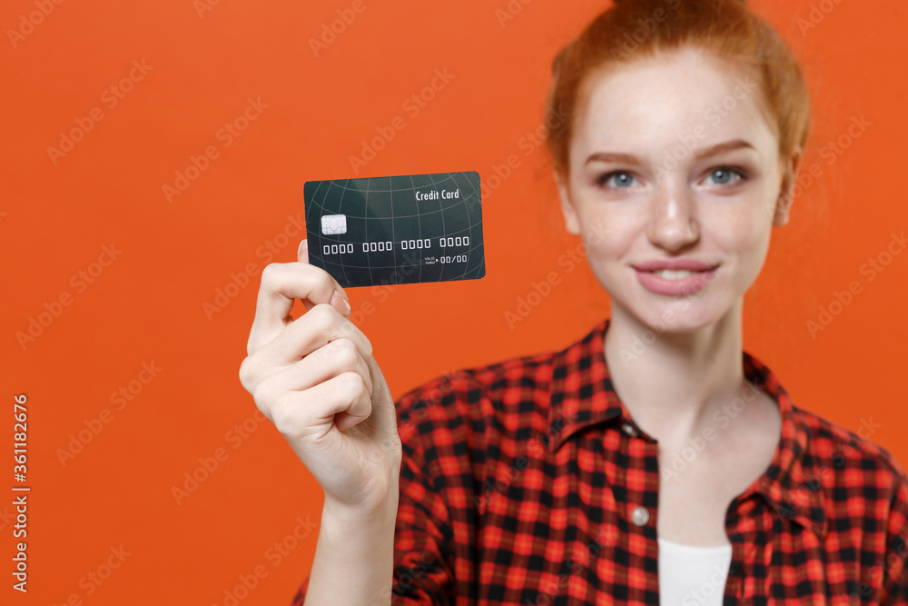Smiling young readhead girl in casual red checkered shirt posing isolated on orange background studio portrait. People sincere emotions lifestyle concept. Mock up copy space. Hold credit bank card. Smiling young readhead girl in casual red checkered shirt posing isolated on orange background studio portrait. People sincere emotions lifestyle concept. Mock up copy space. Hold credit bank card.
